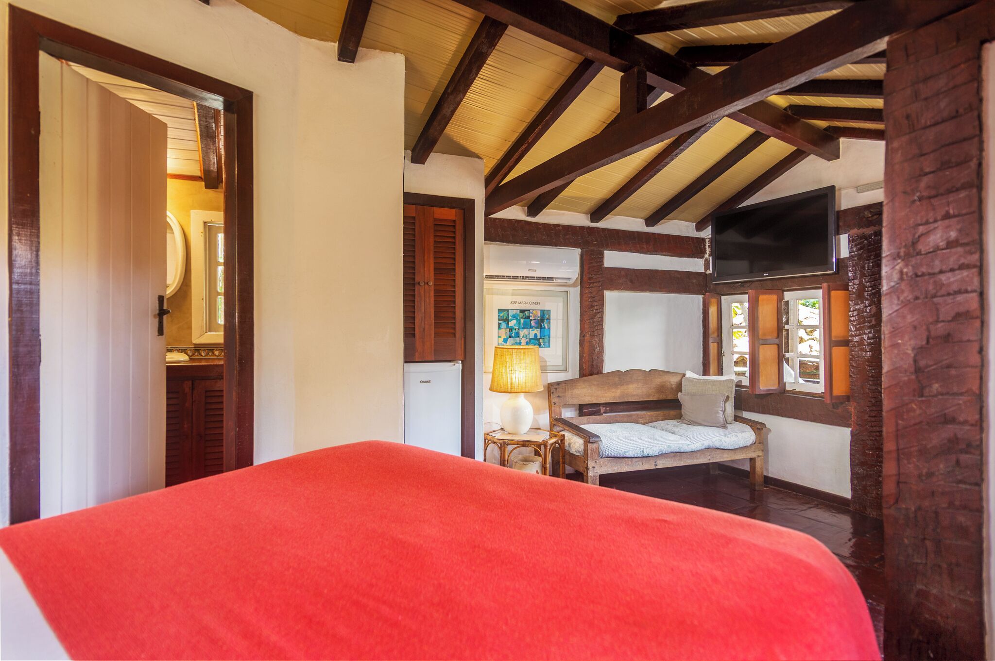 A bedroom with a wooden ceiling, a bed with a red cover, a wooden bench, a lamp, and a television on the wall.