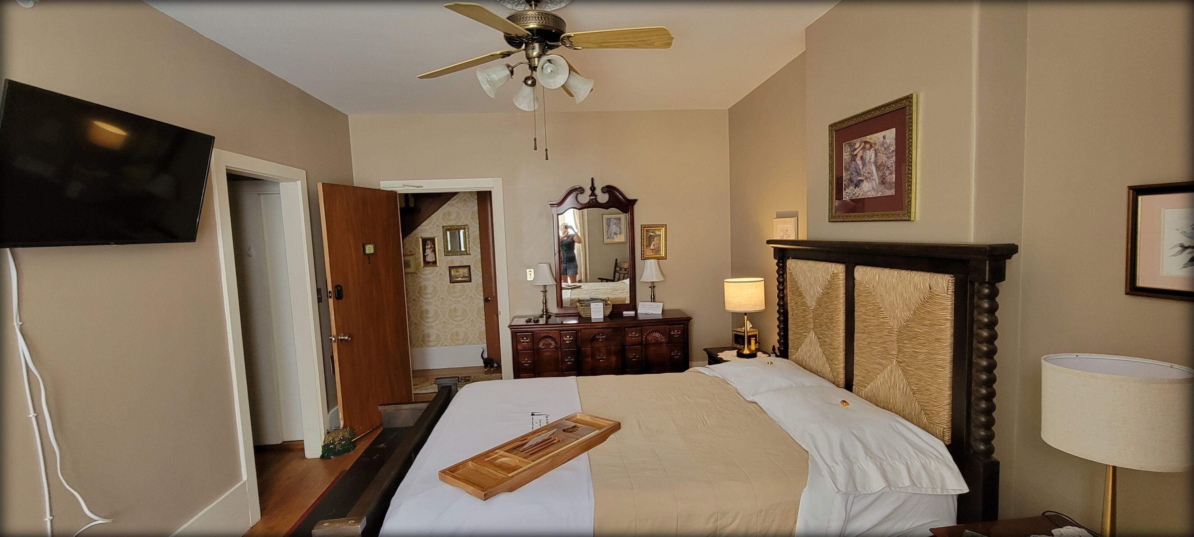 Une chambre d’hôtel avec un lit, une table de chevet en bois, une lampe, un téléviseur et un ventilateur de plafond.