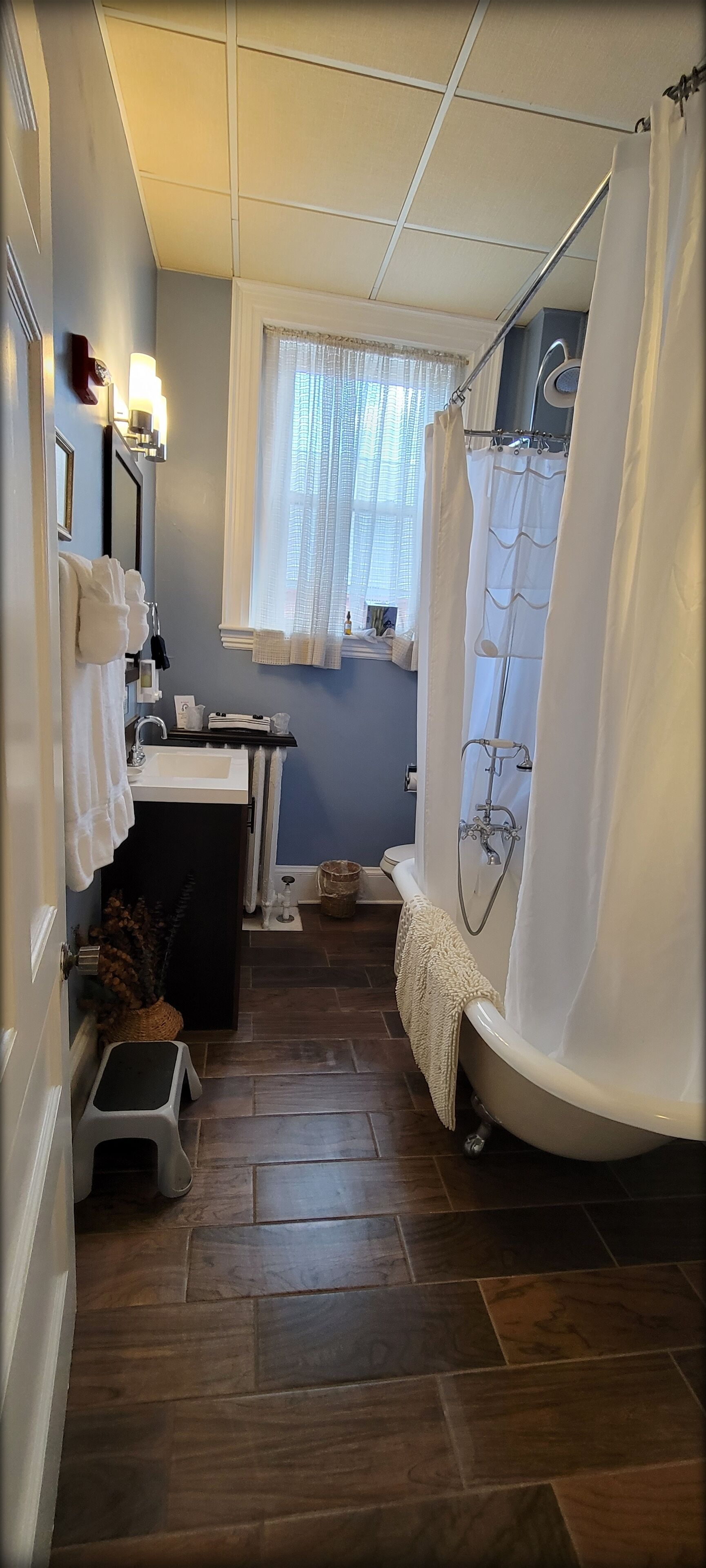 A narrow bathroom with a bathtub, a sink, and a window with sheer curtains.