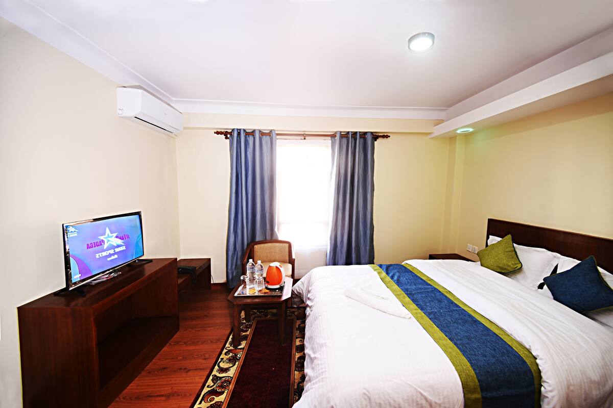 A hotel room with a bed, a television on a stand, a desk with a chair, and a window with curtains.