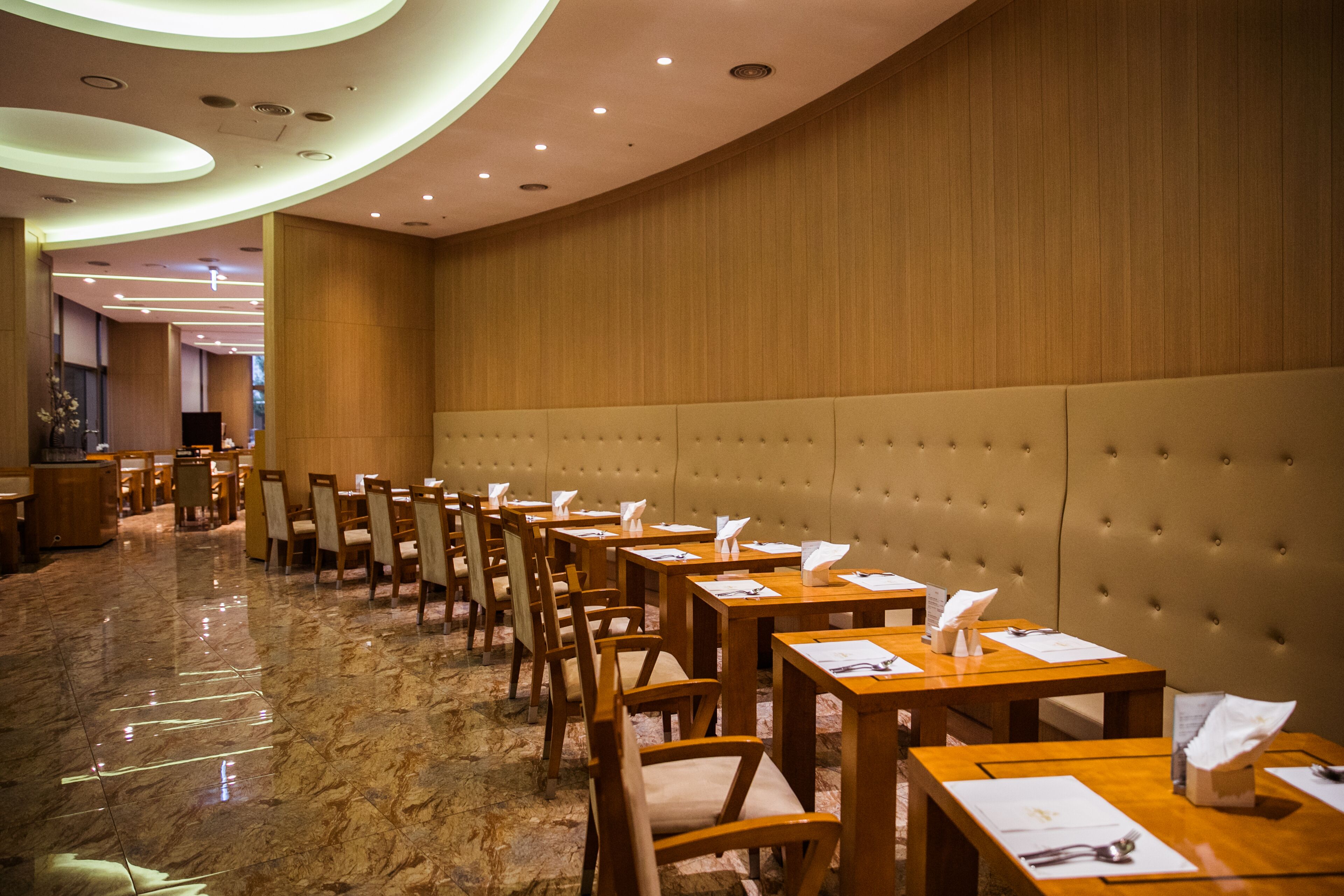 A hotel lobby with a row of wooden tables and chairs, a marble floor, and a tufted wall.