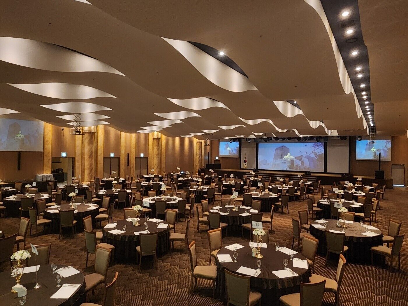 Une salle de conférence dotée de plusieurs tables et chaises, équipée d’écrans de projection et d’une scène.