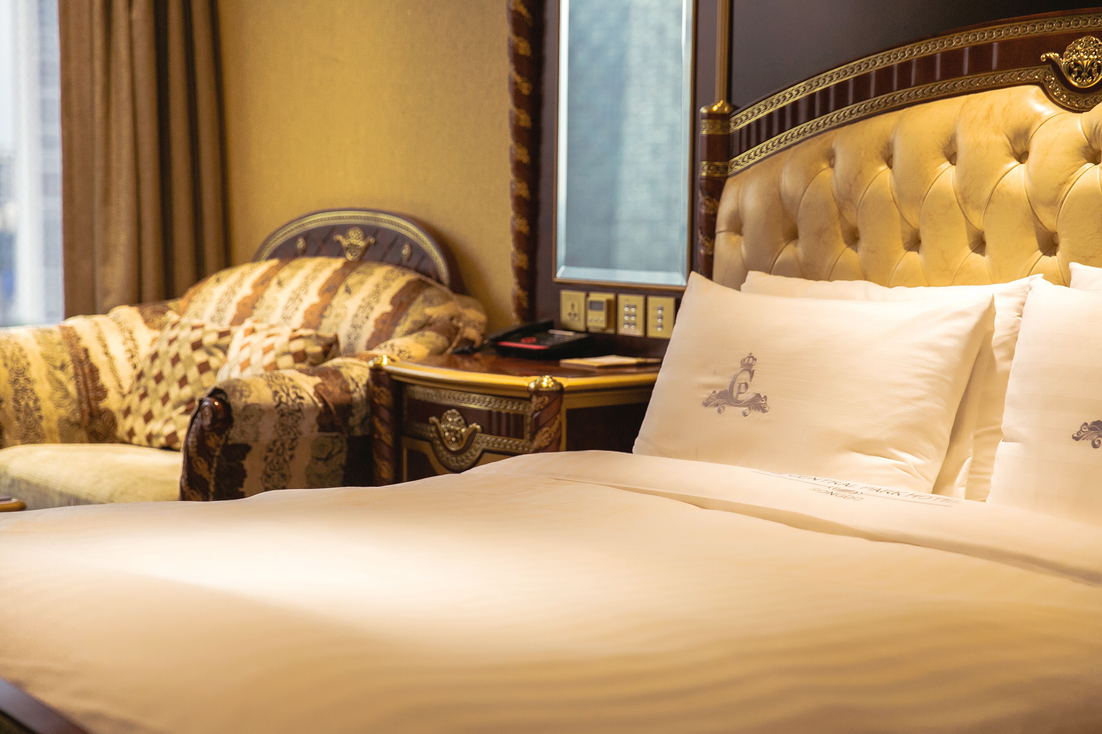 A luxurious hotel room with a large bed, a tufted headboard, a classic armchair, and a bedside table with a lamp.