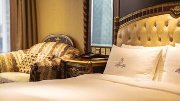 A luxurious hotel room with a large bed, a tufted headboard, a classic armchair, and a bedside table with a lamp.