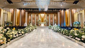 Un amplio salón con suelo reflectante, flanqueado por arreglos florales y columnas, que conduce a un escenario con un atril.