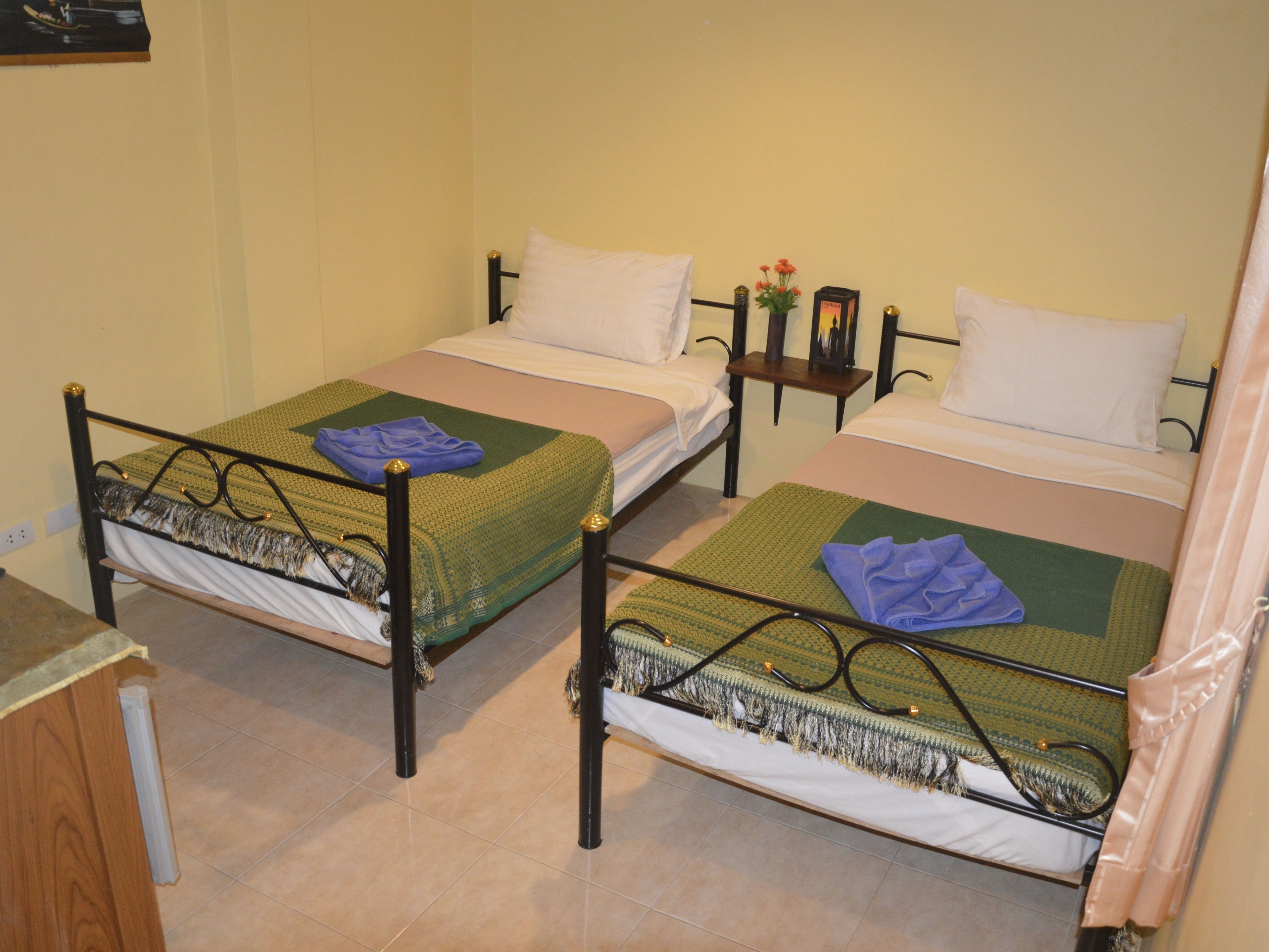Una habitación con dos camas individuales, cada una con una colcha verde y blanca, una mesita de noche con lámpara y un cuadro en la pared.