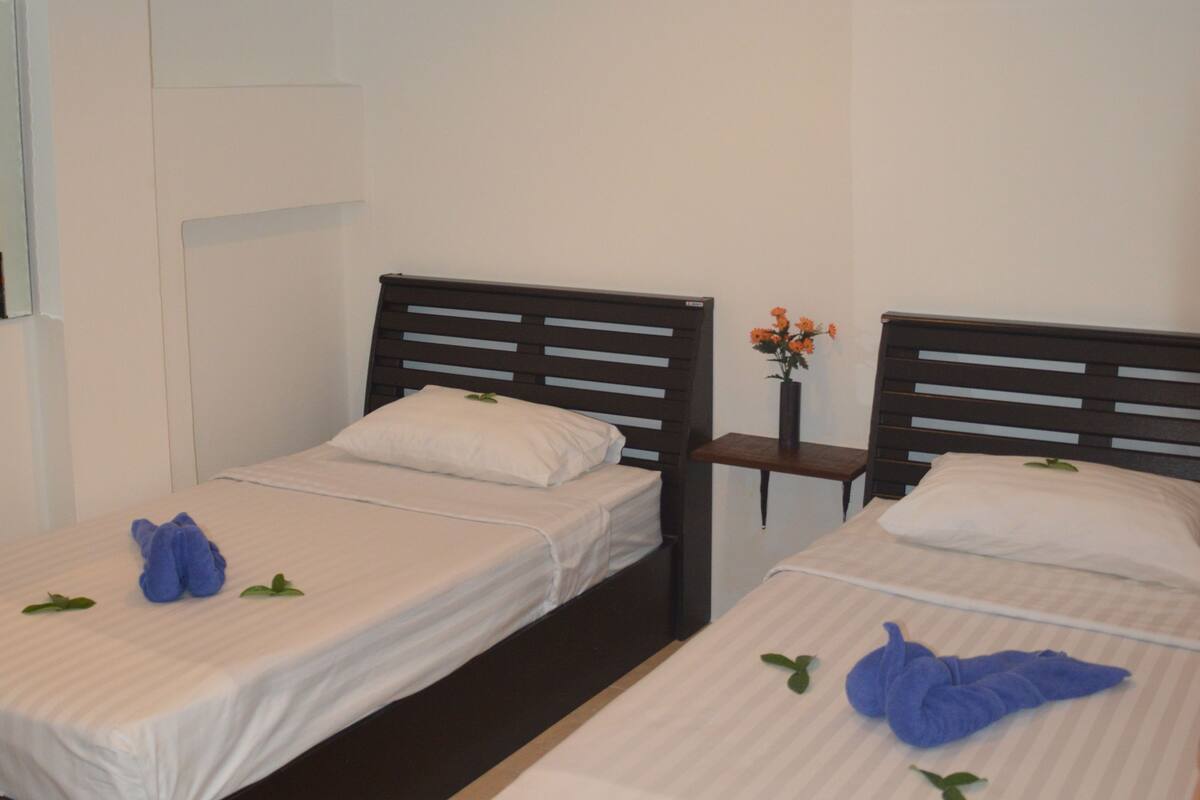 A hotel room with two single beds, each with a wooden headboard, white linens, and a small table with a vase of flowers between them.