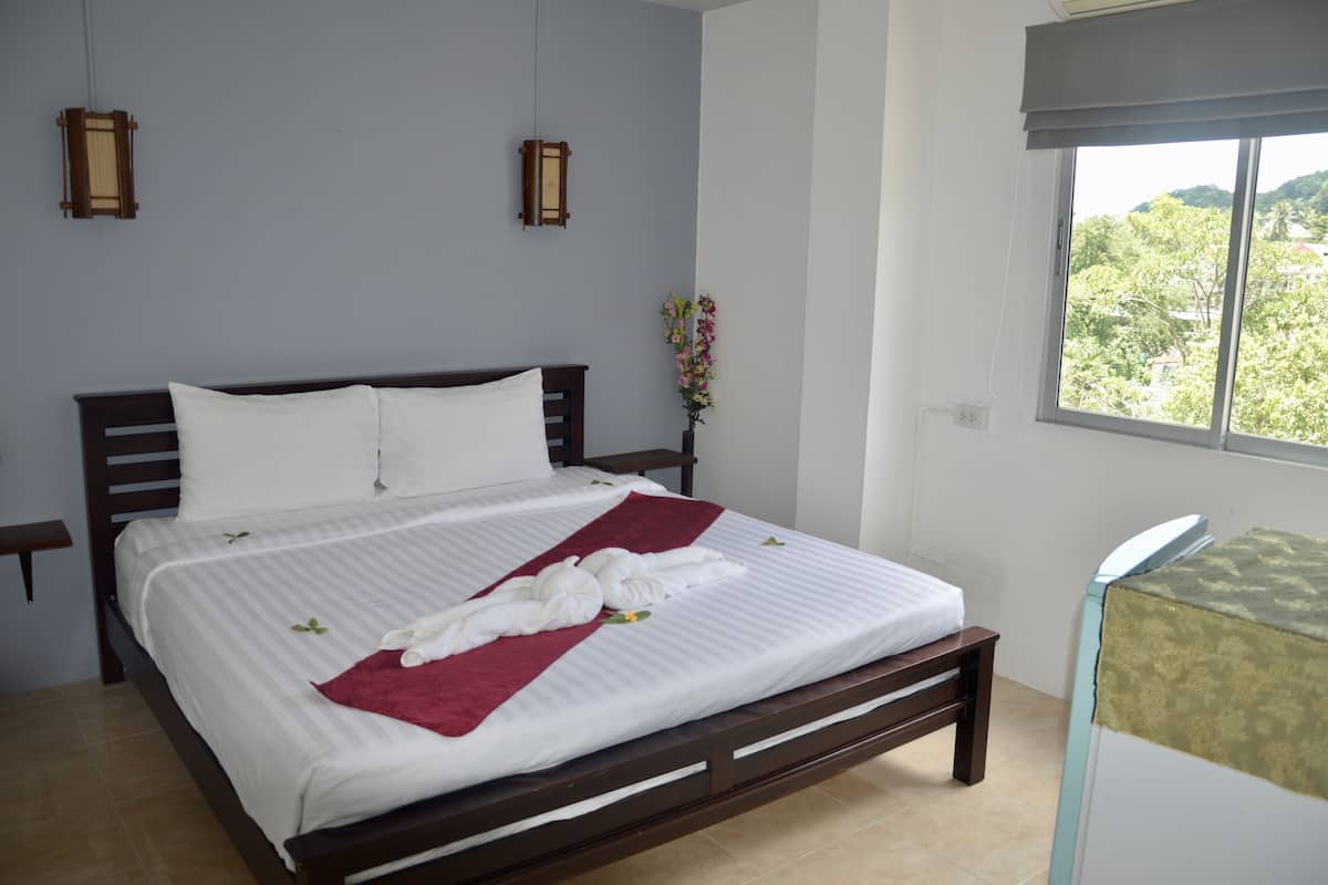 A neatly made bed with white linens and a red throw blanket, a wooden headboard, and a window with a view of greenery.