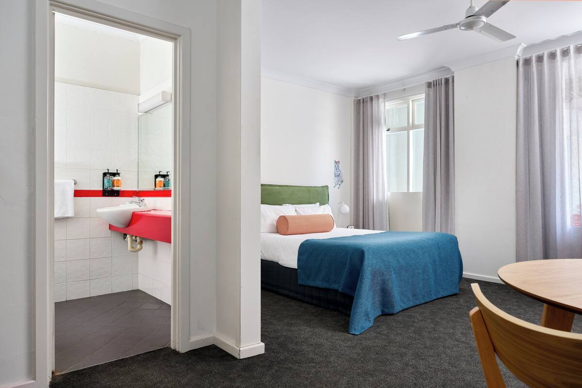 Una camera d'albergo con un letto, un comodino, una sedia, una scrivania, una finestra con tende e un bagno con lavandino e specchio.