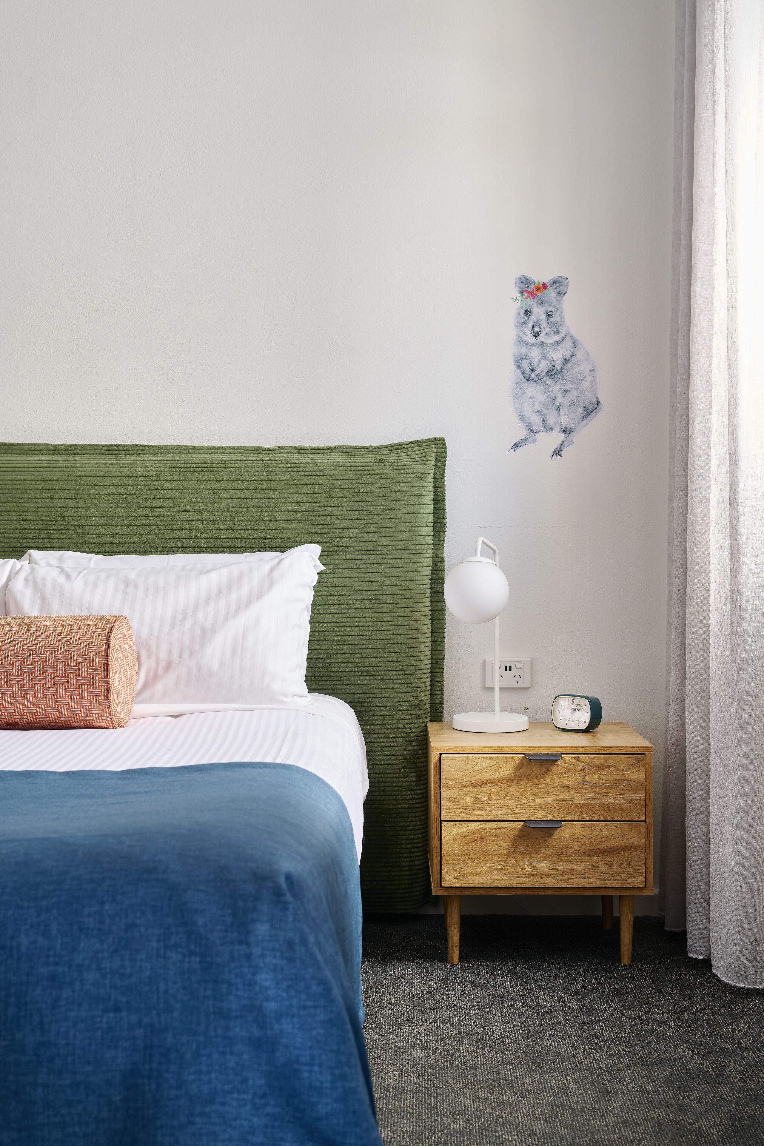 A neatly made bed with a green headboard, a bedside lamp, a small wooden nightstand with a clock, and a wall decal of a koala.