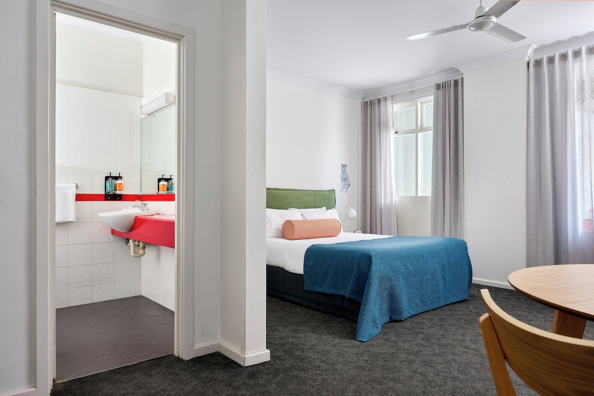 A hotel room with a bed, a desk, a chair, a bathroom with a sink and mirror, and a ceiling fan.
