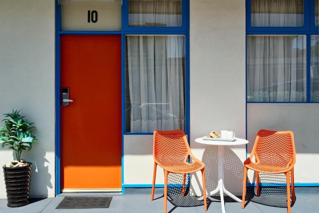A hotel room with a red door, two orange chairs, a small round table, and a potted plant.