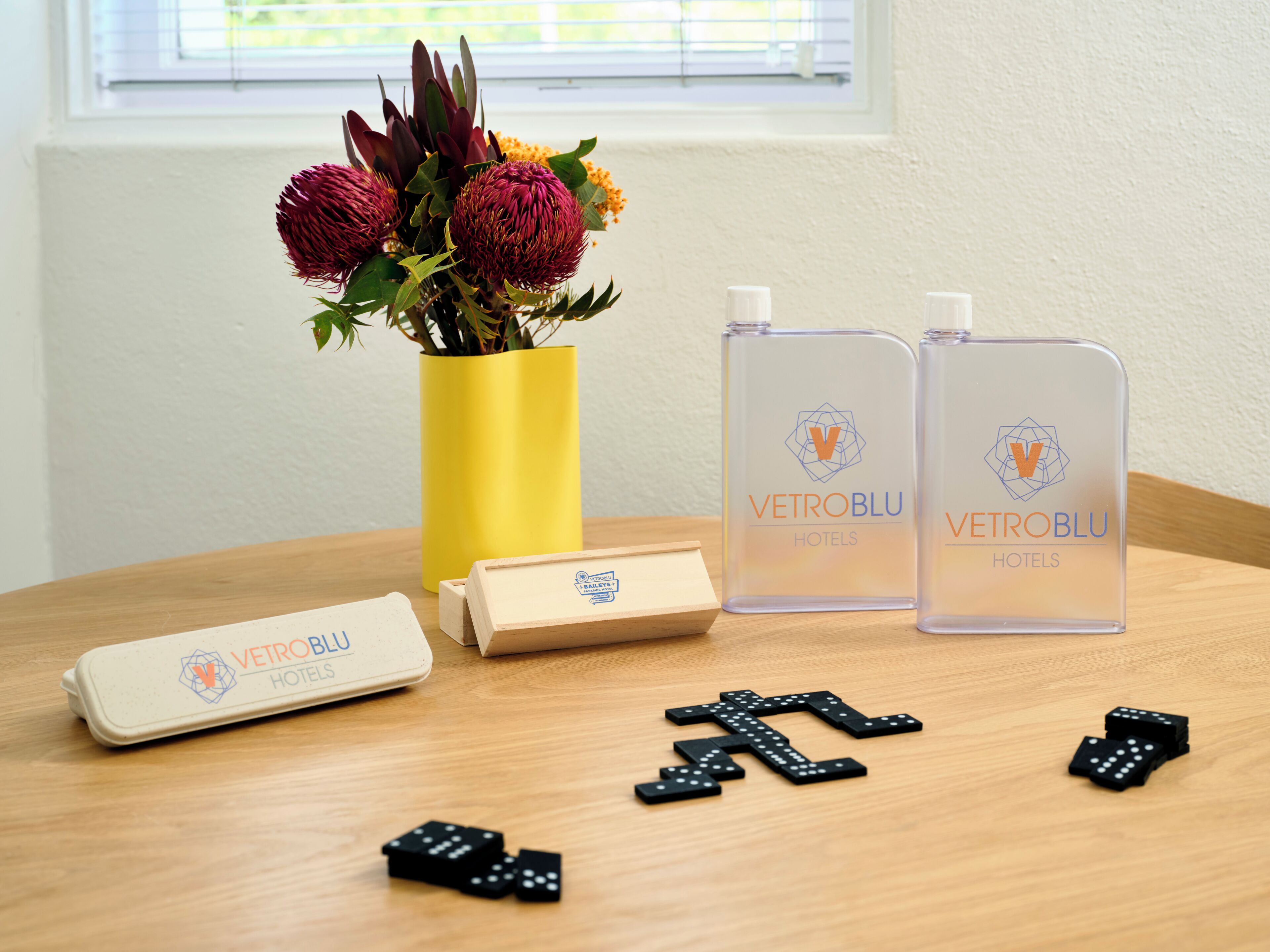 A table with a vase of flowers, two bottles, a box, and dominoes.