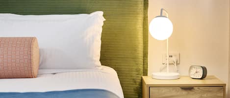 Een net opgemaakt bed met wit- en blauw beddengoed, een groen hoofdeinde, een bedlampje, een klein houten nachtkastje met een klok en een muurstickers van een kangoeroe.