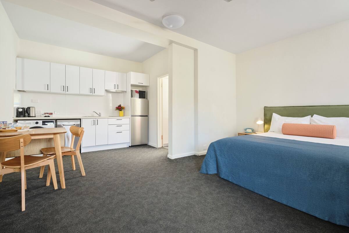 Una camera d'albergo compatta con angolo cottura, un letto con copriletto blu e una ventola a soffitto.