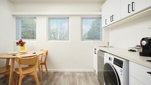A modern kitchen with a built-in washer, a dining area with wooden chairs, and three windows with blinds.