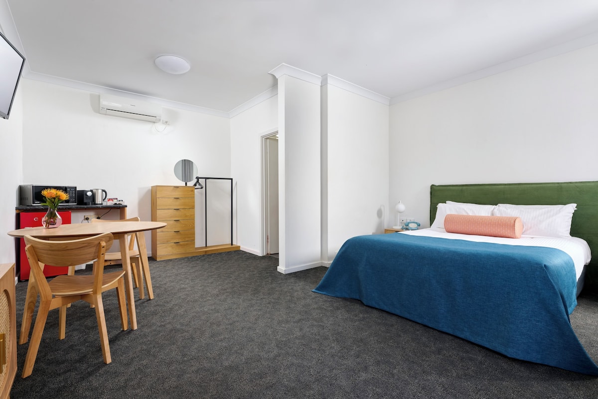 Una camera d'albergo con un letto, una scrivania, una sedia, un comò e un tavolino con un vaso di fiori.