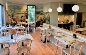 A restaurant with round tables and chairs, a mural on the wall, and pendant lighting. - Baileys Motel (East Perth)