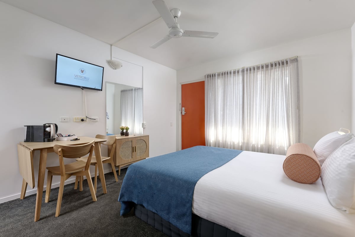 Una camera d'albergo con un letto, una scrivania con una sedia, una televisione e una ventola a soffitto.
