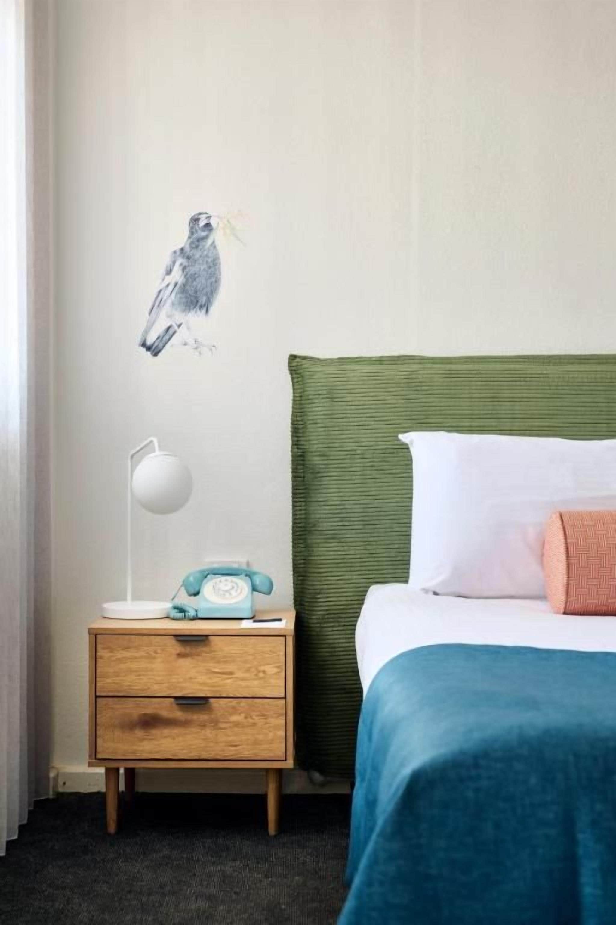 A hotel room with a bed, a nightstand with a lamp, a telephone, and a wall-mounted artwork.