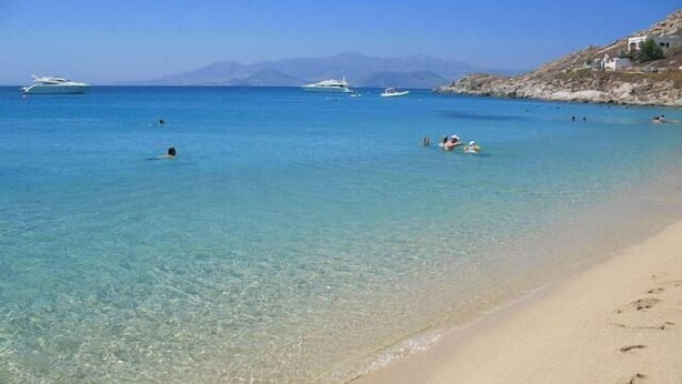 A beach with clear turquoise water, a sandy shore, and boats in the distance. - Kahlua Studios (Naxos)