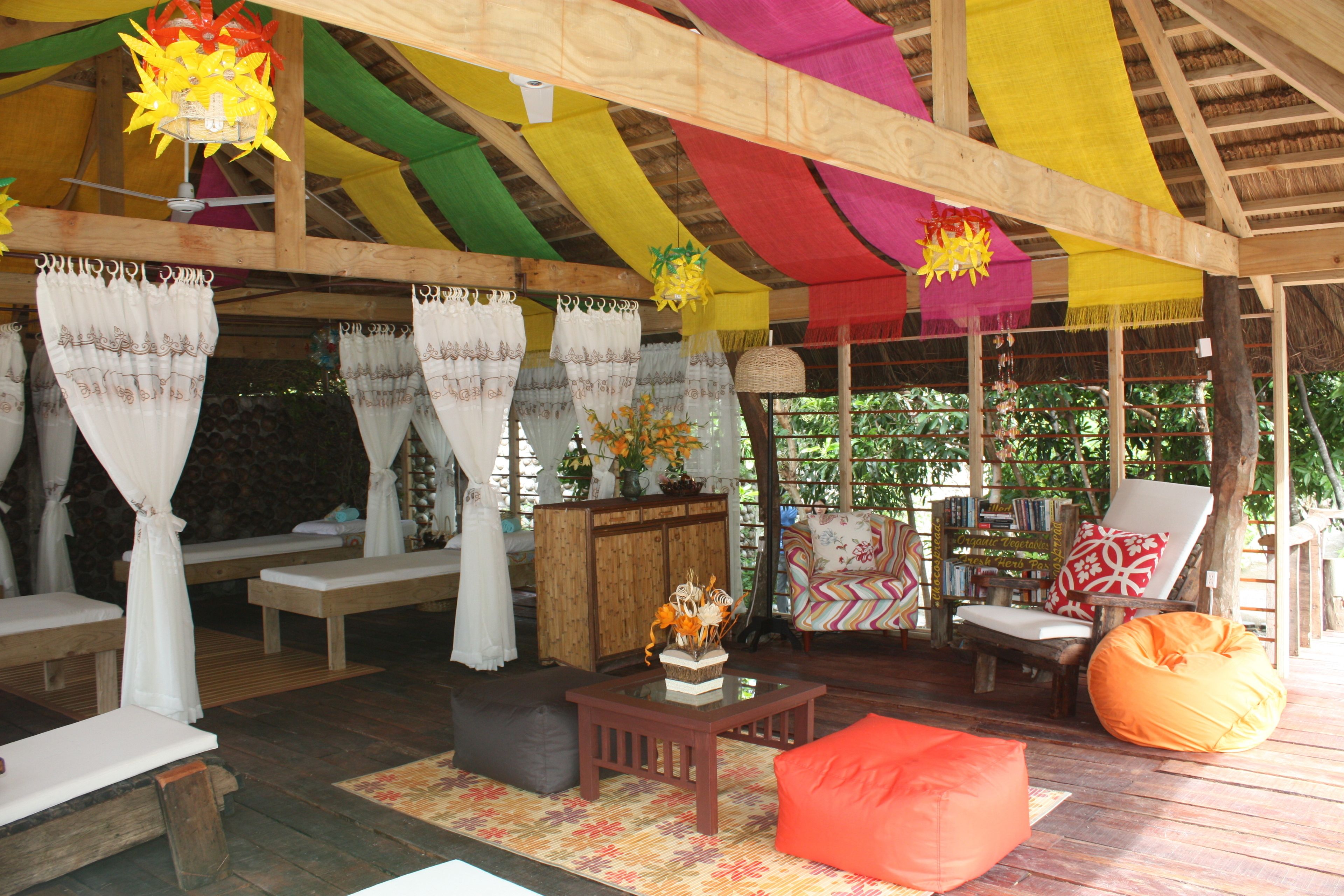 A covered outdoor seating area with wooden furniture, a table with a vase, and colorful fabric drapes.