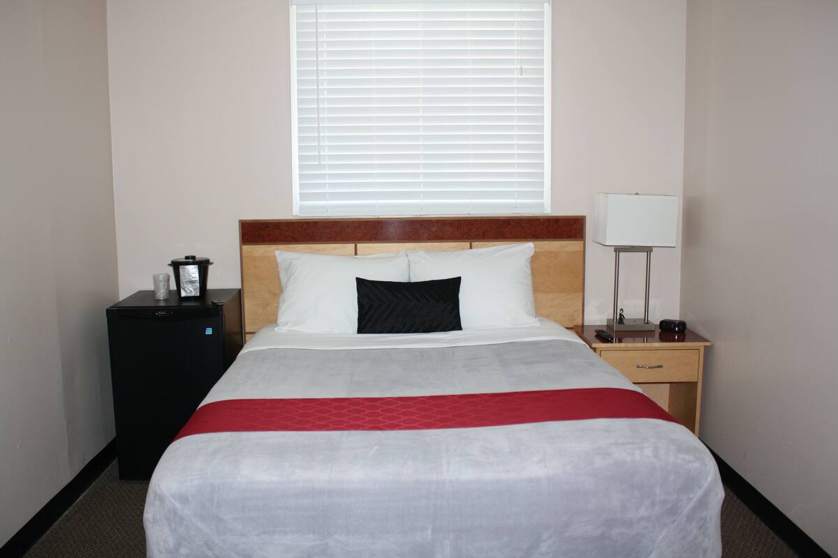 A hotel room with a bed, a nightstand, a lamp, a heater, and a window with blinds.