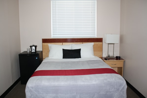 A hotel room with a bed, a nightstand, a lamp, a heater, and a window with blinds. - Bexon Rooms - Hotel Downtown Windsor (Windsor)