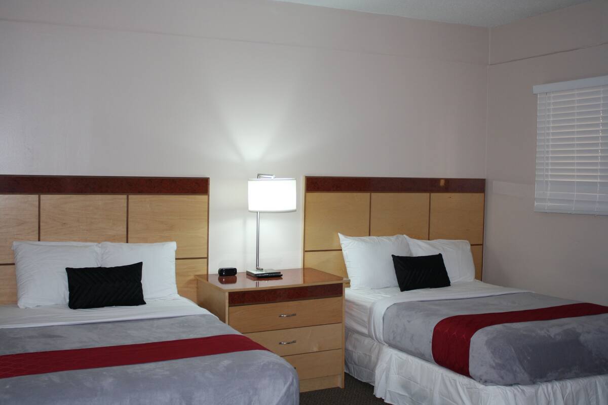 A hotel room with two beds, a bedside table with a lamp, and a window with blinds.