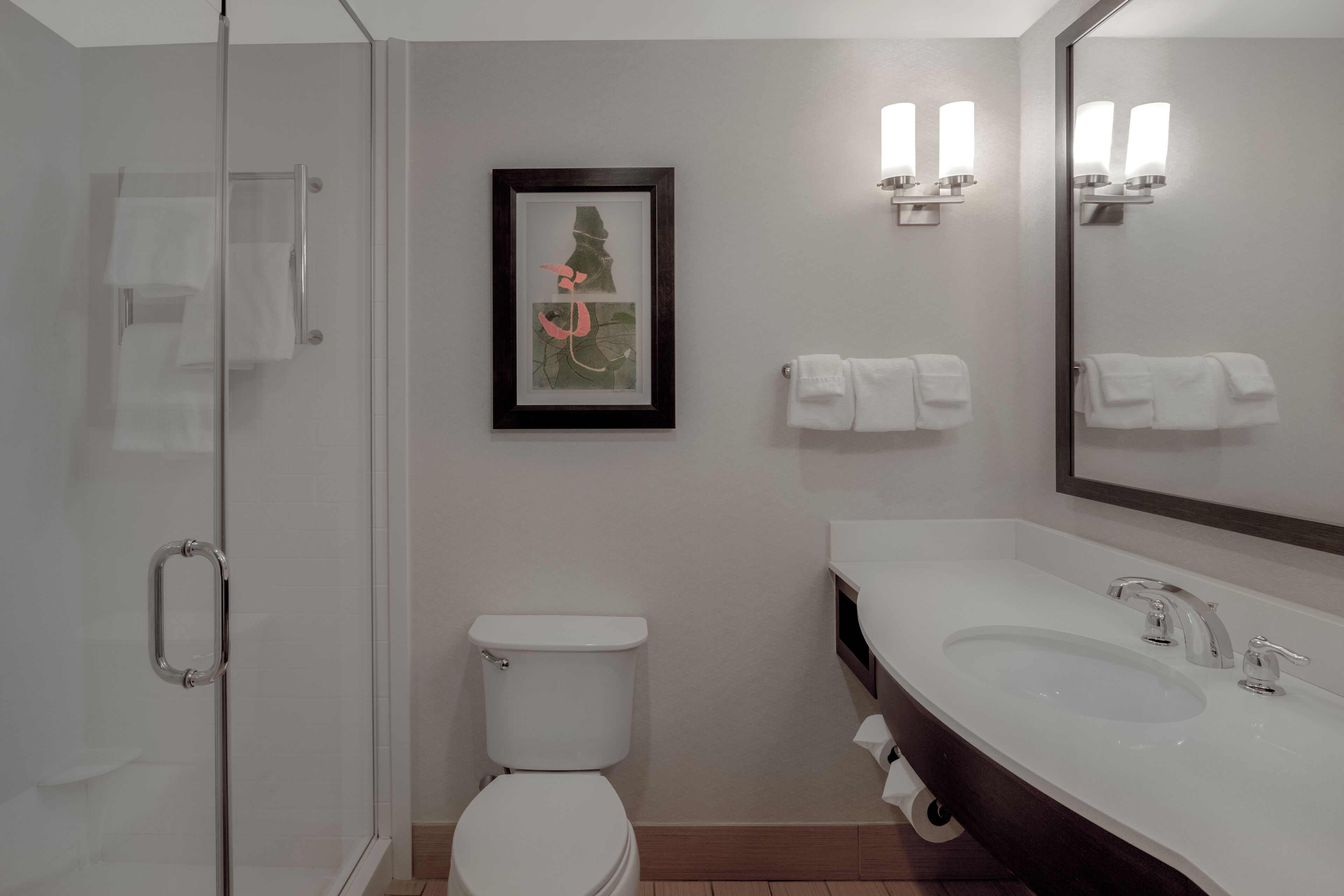 A modern bathroom with a glass shower enclosure, a white toilet, a large sink with dual faucets, and a framed artwork on the wall.