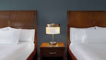 Two neatly made beds with white linens, a wooden nightstand with a lamp between them, and a dark blue wall.