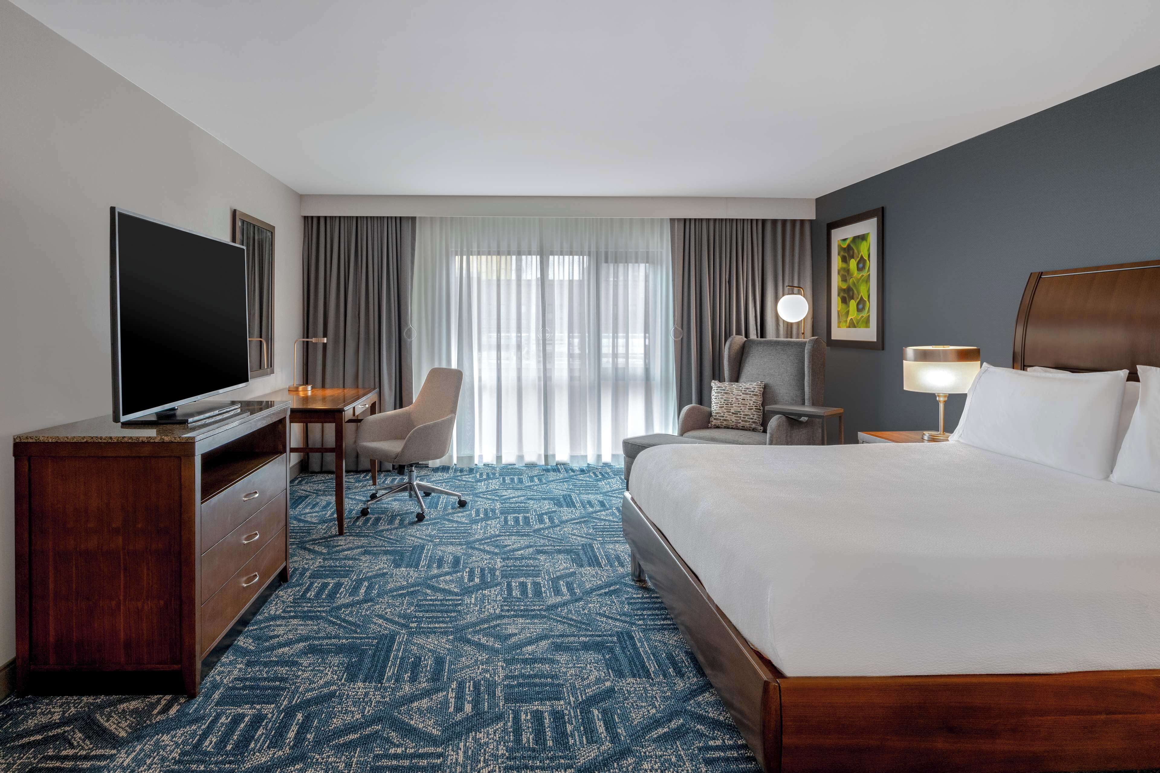 Une chambre d’hôtel avec un grand lit, un bureau avec une chaise, une télévision et une fenêtre avec des rideaux.
