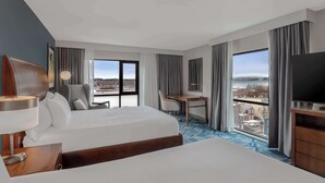 A hotel room with two beds, a desk, a chair, a TV, and large windows offering a city view.