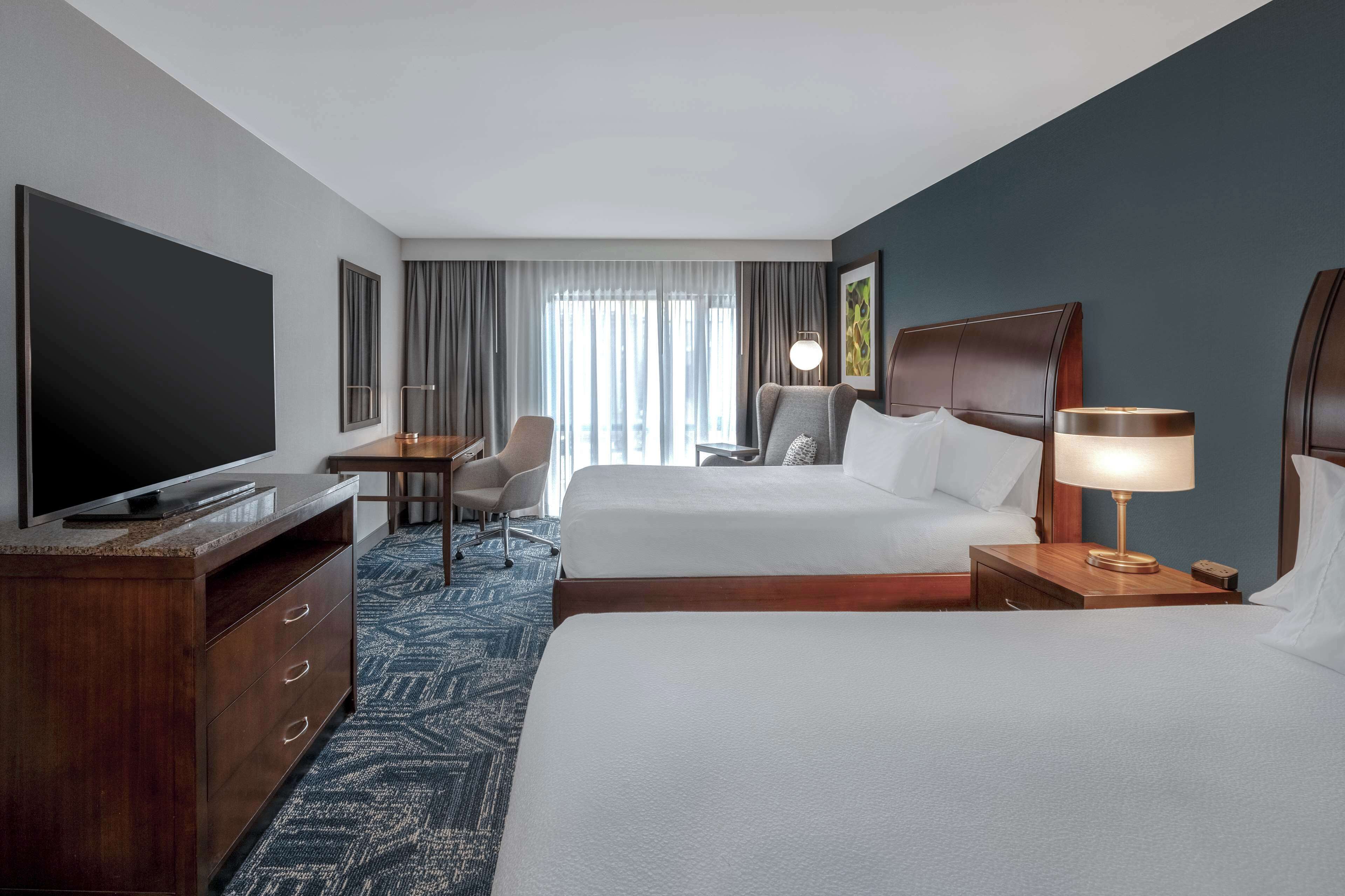 Quarto de hotel com uma televisão de tela grande, duas camas, uma escrivaninha e uma cadeira.