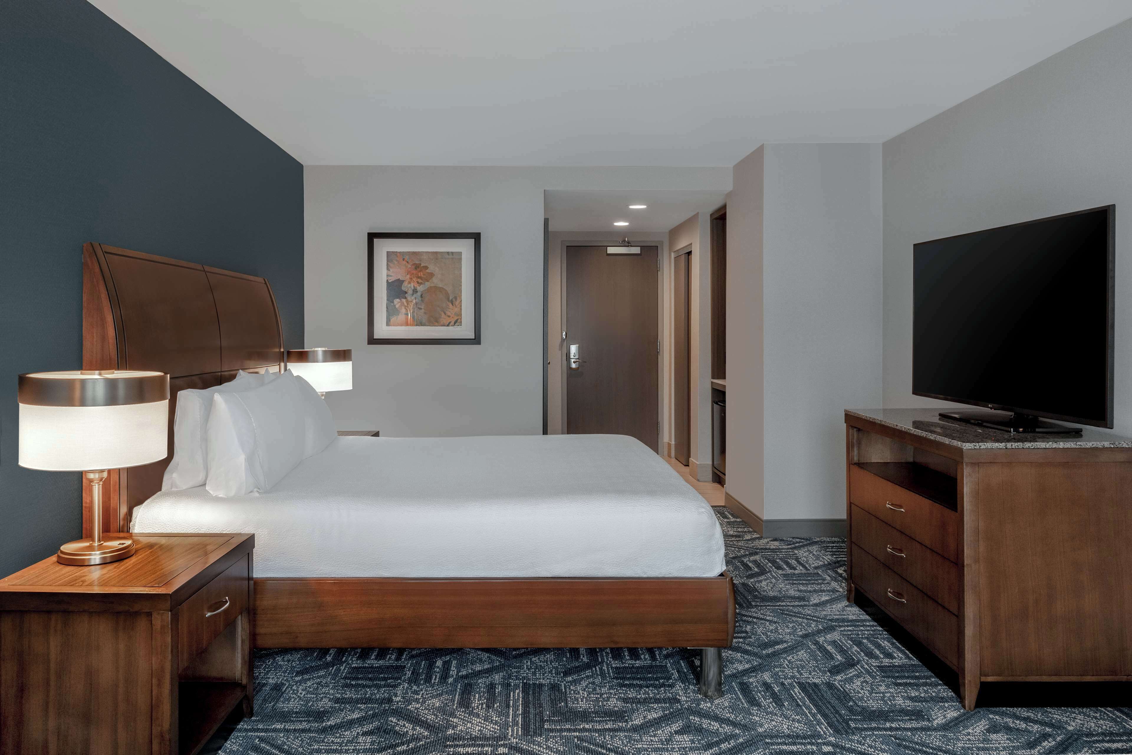 Een hotelkamer met een bed, een televisie, een bureau met een lamp en een schilderij aan de muur.