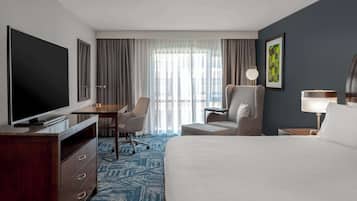 A hotel room with a large bed, a desk with a chair, a television, a lamp, and a window with curtains.