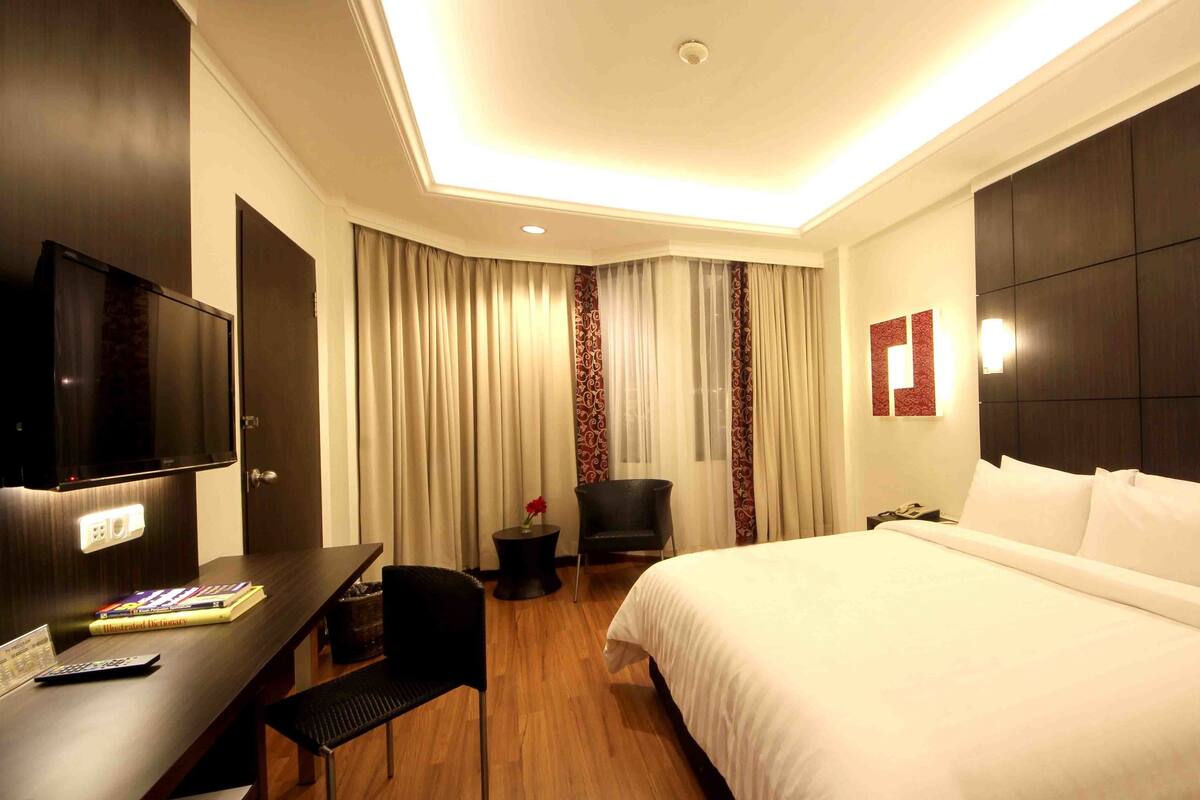 A hotel room with a large bed, a TV mounted on the wall, a desk with a chair, and a small table with a vase of flowers.