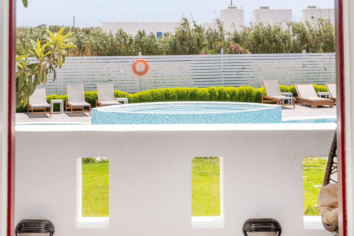 A view through an arched doorway of a pool area with lounge chairs, a clock, and a building in the background.