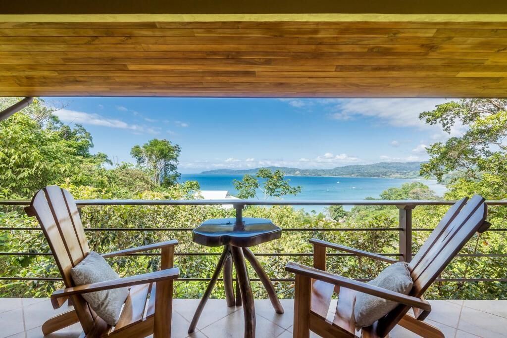 A balcony with two wooden chairs and a small table, overlooking a scenic landscape with trees and a view of the sea.