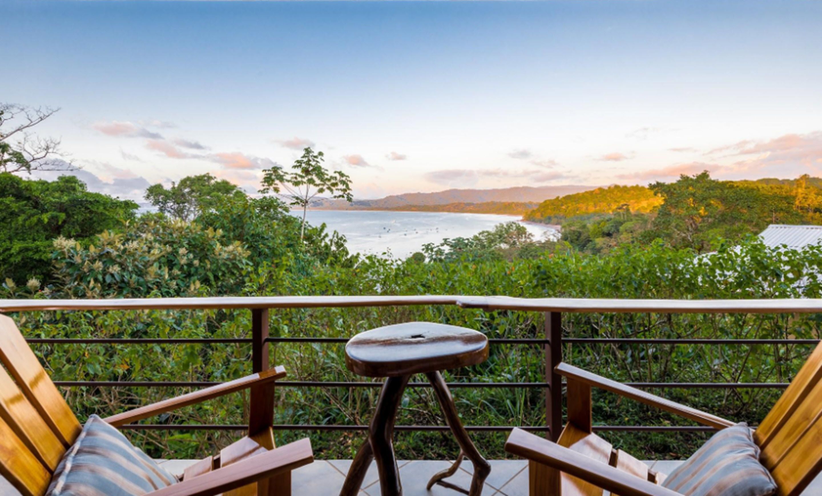 A balcony with two wooden chairs and a small table, overlooking a body of water and lush greenery.