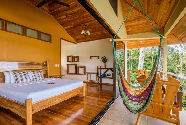 A bedroom with a wooden bed, a hammock, and a balcony with a view of trees.