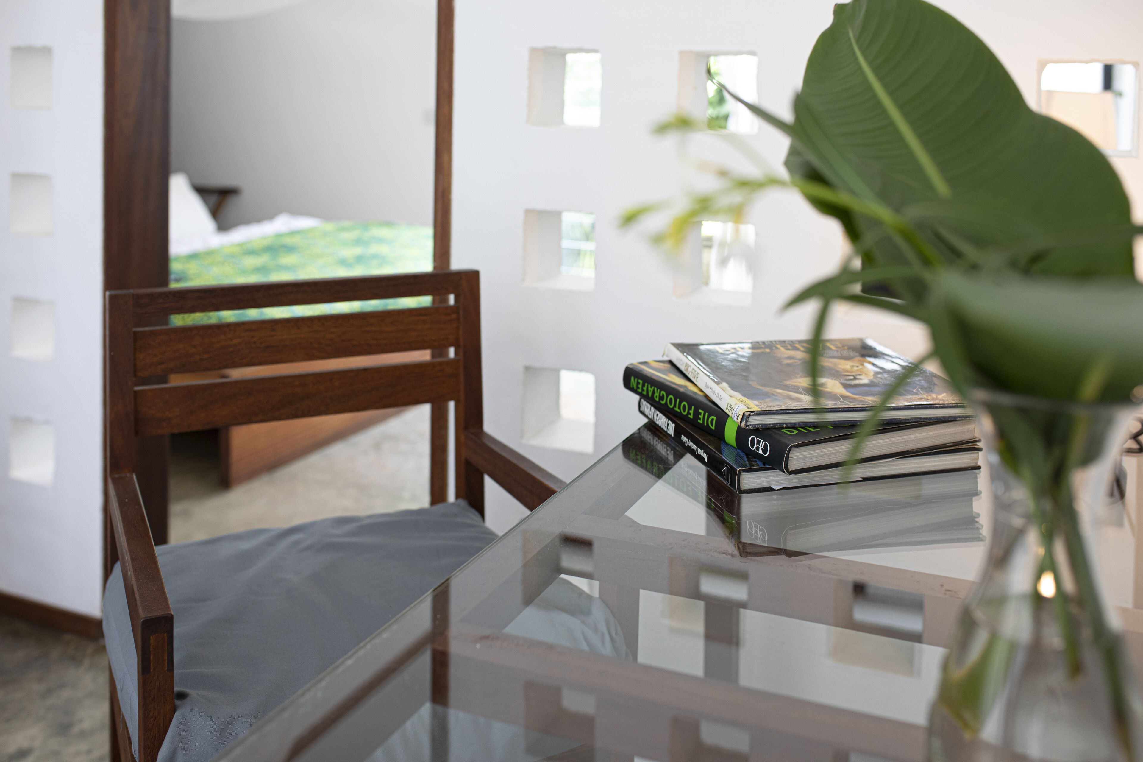 A room with a bed, a wooden chair, a glass table with books, and a plant.