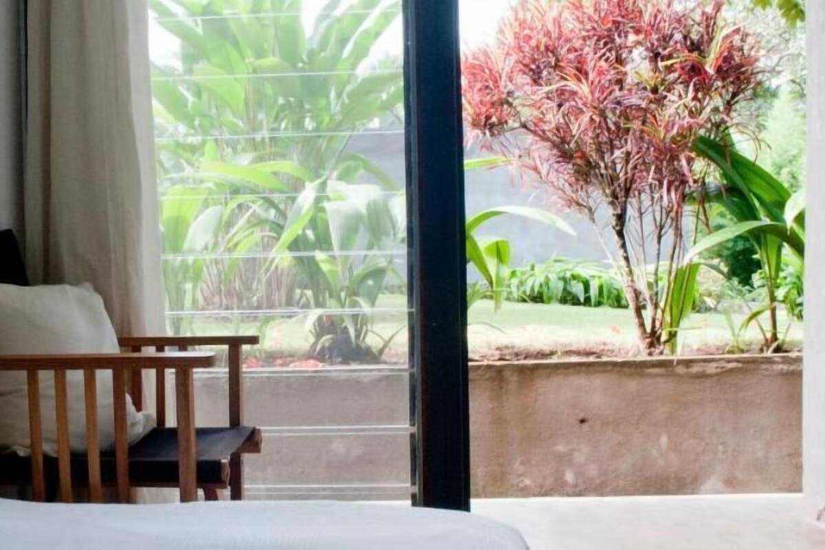 A room with a bed, a wooden bench, a window with a view of greenery.