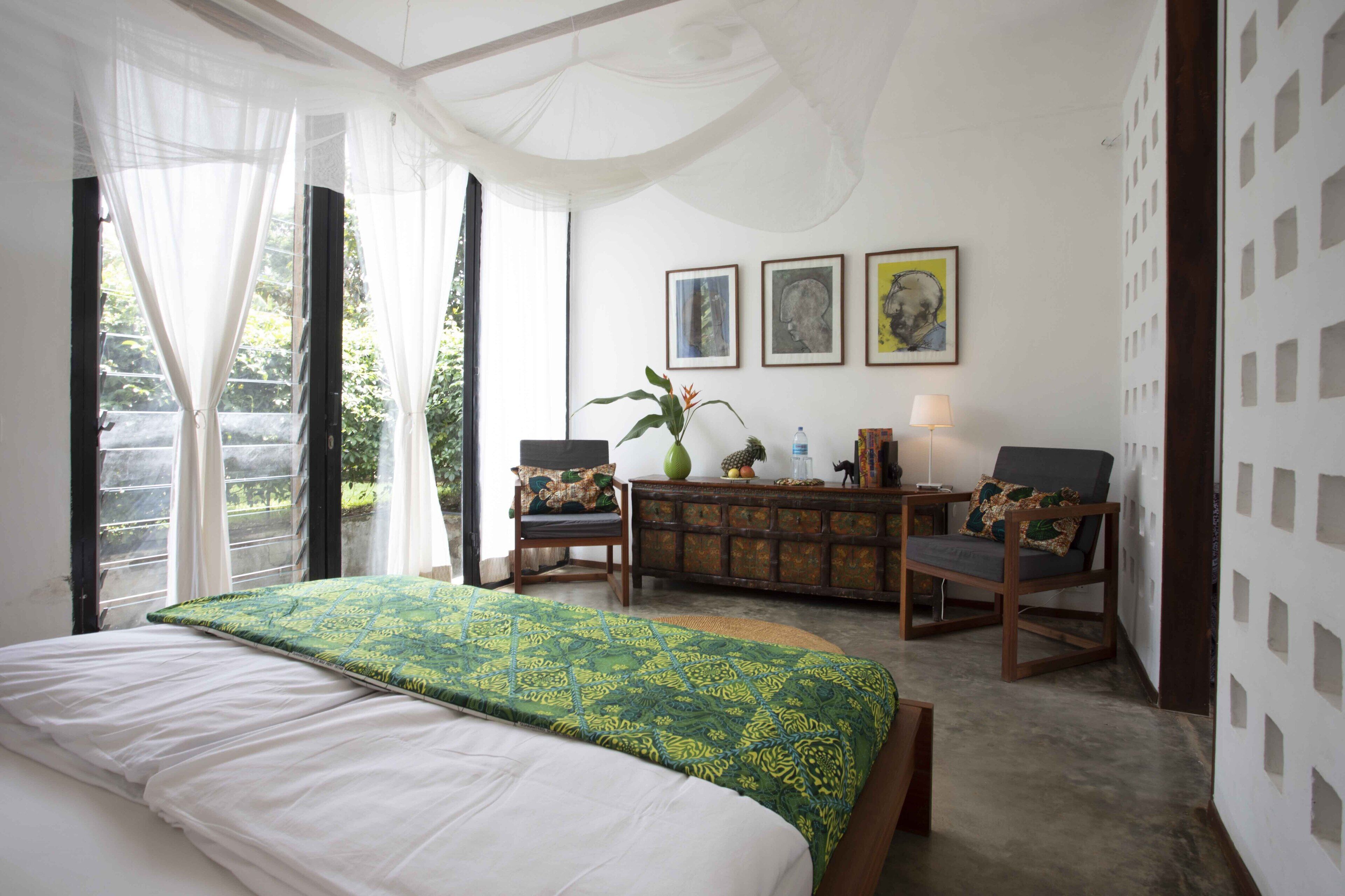 Un dormitorio con una cama grande, un escritorio, dos sillas, un florero y obras de arte enmarcadas en la pared.