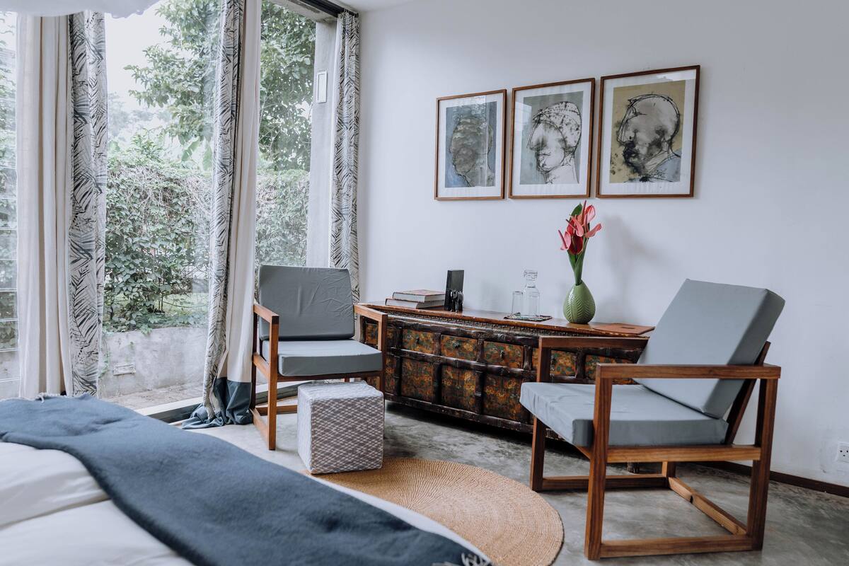 A modern bedroom with a bed, two chairs, a small table, a decorative vase, and framed artwork on the wall.