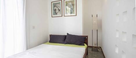 A bedroom with a bed, two framed pictures on the wall, a floor lamp, and a white wall with square cutouts.