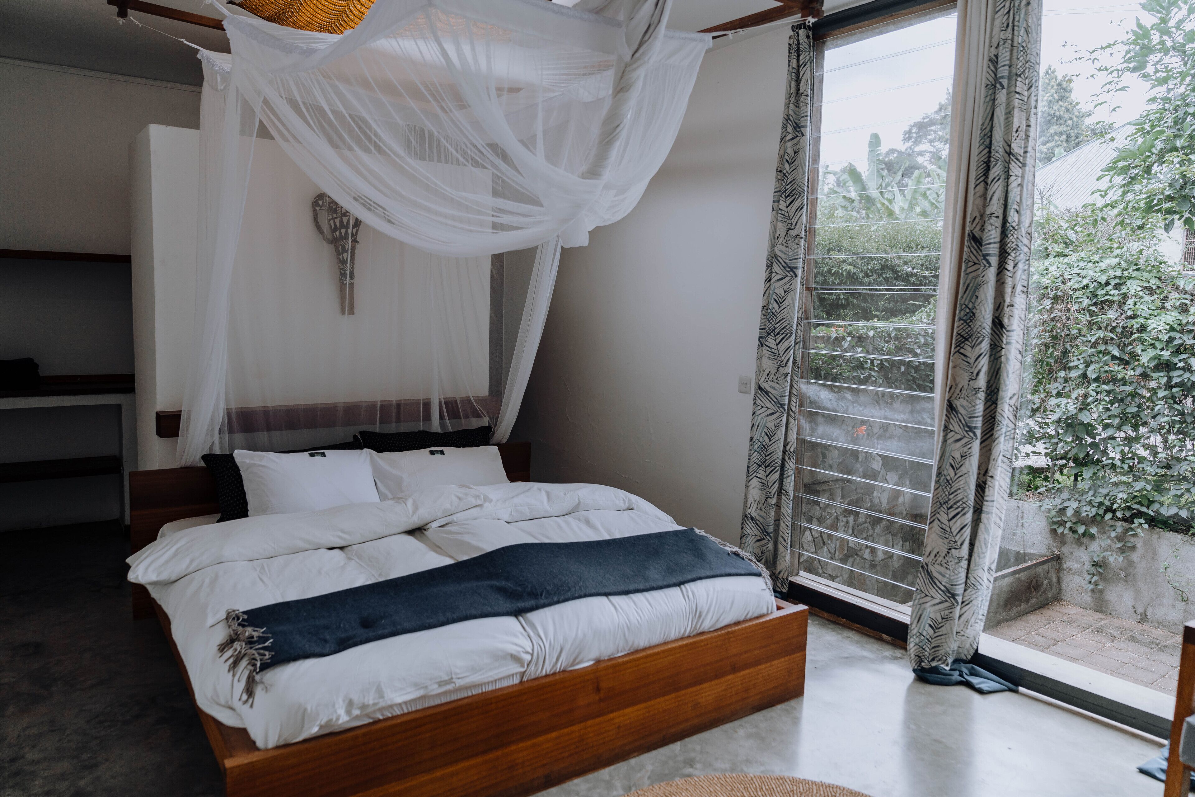 A bedroom with a wooden bed, white bedding, a canopy, and a view of greenery outside.