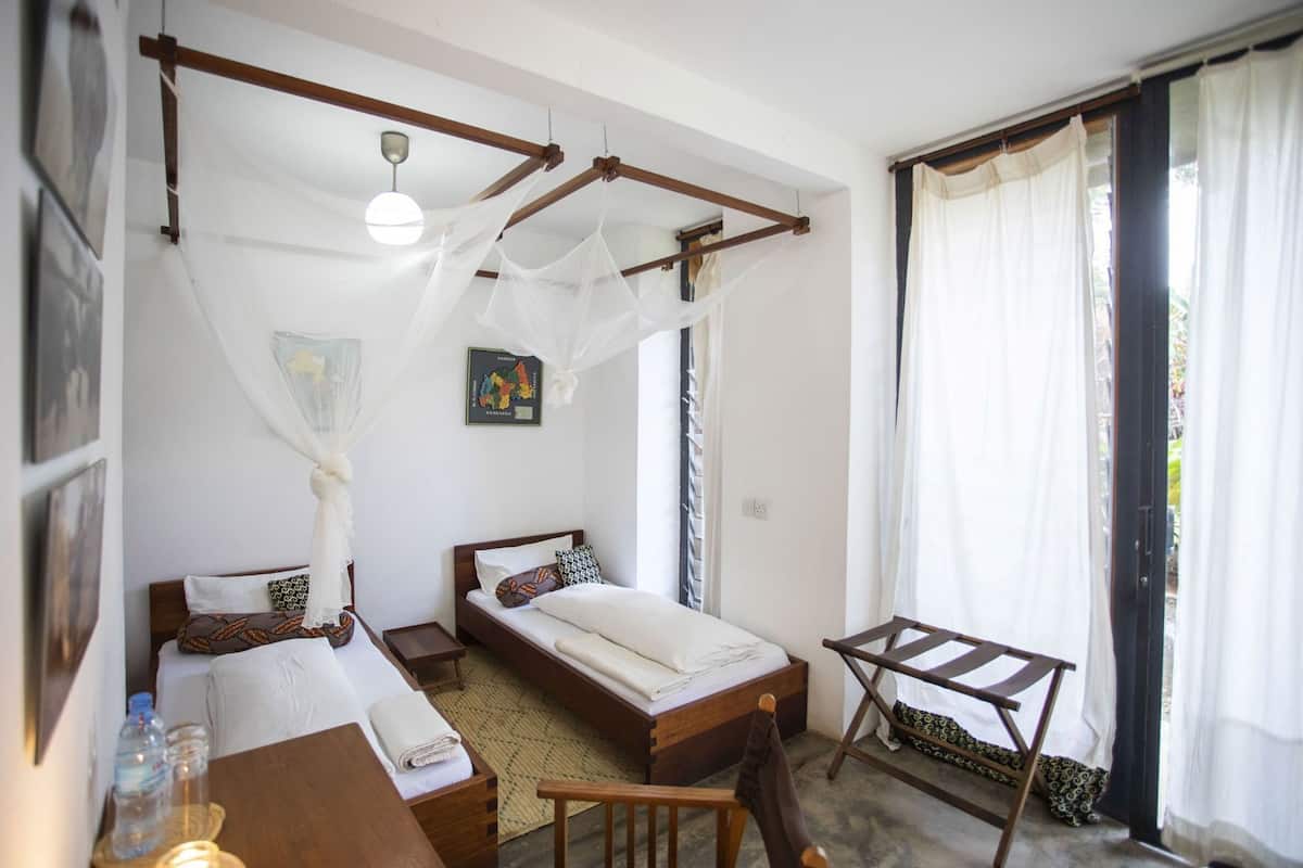 A bedroom with two beds, a wooden chair, a table, a map on the wall, and a window with curtains.