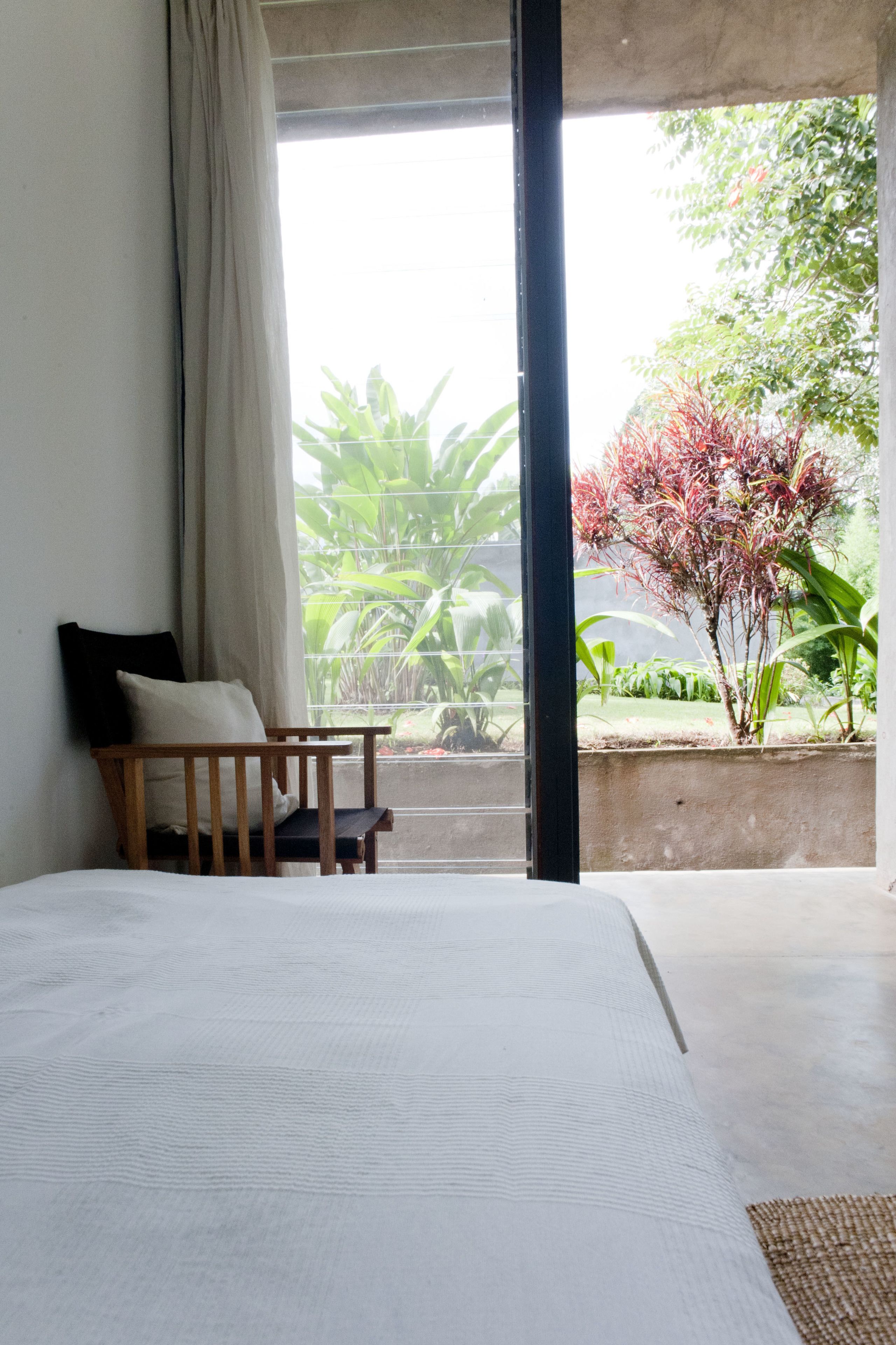 A bedroom with a large bed, a wooden chair, a window with a view of greenery and a tree.