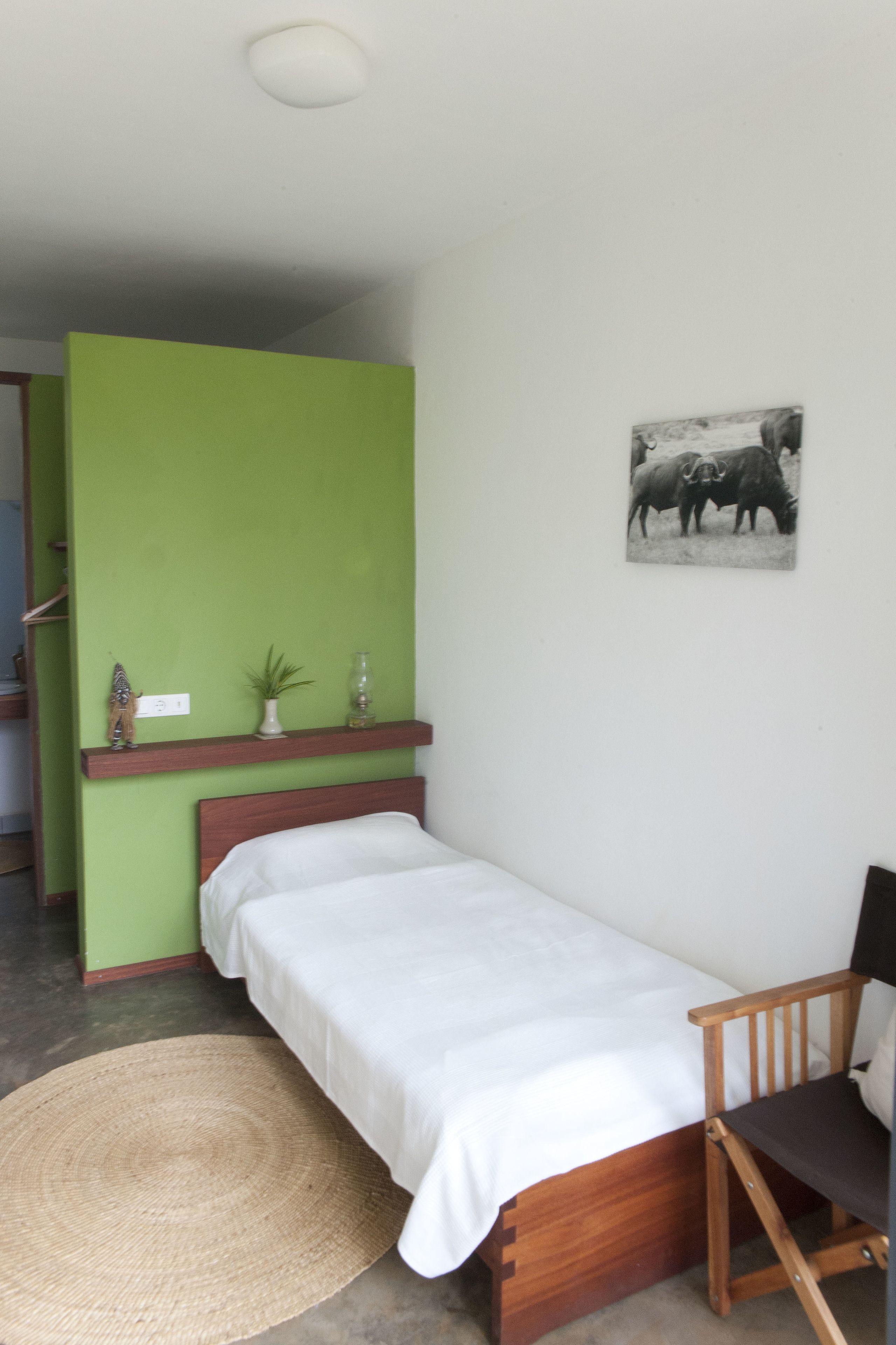 A small, single-bed room with a green wall, a wooden chair, a small shelf with decorative items, and a picture of bulls on the wall.